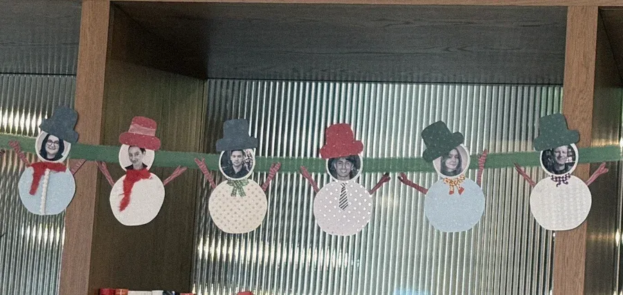 A paper snowman garland strung across a bookshelf, each snowman with a teammate’s face