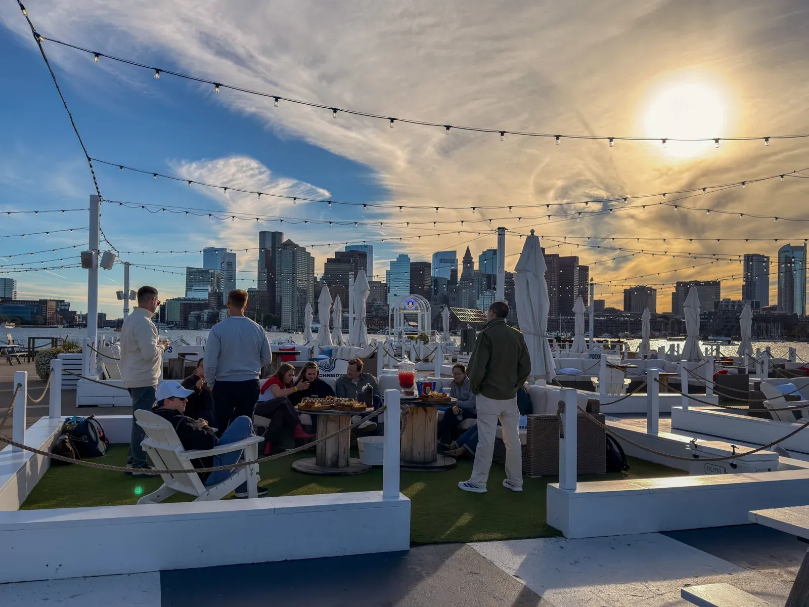 The design team relaxing on the tall ship’s deck under string lights, with the Boston skyline at sunset behind them